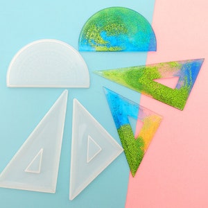 Puede incluir: Cinco formas geométricas de plástico transparente, incluyendo dos triángulos, dos semicírculos y un triángulo con un diseño de purpurina azul, amarillo y verde.