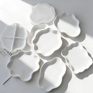 Pode incluir: Um conjunto de pratos de cerâmica brancos com bordas onduladas. Alguns são sólidos, outros têm divisões internas. Os pratos estão dispostos sobre uma superfície branca, com sombras de luz natural. Tema: decoração e utensílios de mesa.