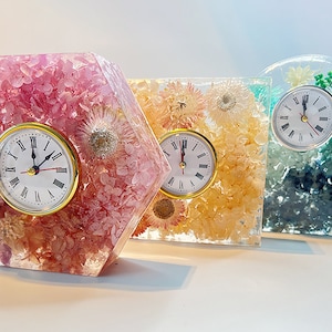 May include: Three resin clocks with embedded dried flowers. The clocks are hexagonal, square, and arched in shape. The flowers are pink, yellow, and blue.