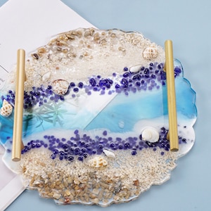 May include: A round resin tray with a beach theme. The tray features a blue and white wave design with white sand, seashells, and purple glass beads. The tray has two gold metal handles.