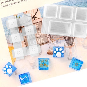 May include: A clear silicone mold with six rectangular cavities for making resin crafts. The mold is on a white surface with several small blue resin pieces with different designs, including a paw print, a gear, and a number.
