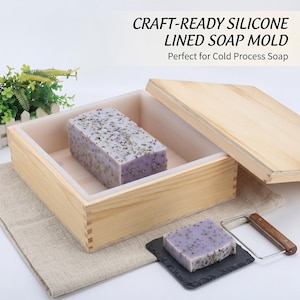 May include: A wooden soap mold with a lavender-colored soap loaf inside. The mold is open, revealing a white silicone lining. A smaller soap bar sits on a black slate, with a soap cutter nearby. Text on the image reads "CRAFT-READY SILICONE LINED SOAP MOLD".