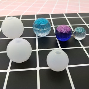May include: Six clear glass balls with glitter inside. The balls are of different sizes and colours, including blue, purple, and white.