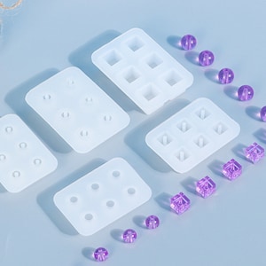 May include: Four silicone moulds for making resin jewellery. The moulds are rectangular and have different numbers of square and round cavities. There are also several purple resin beads and cubes scattered around the moulds.