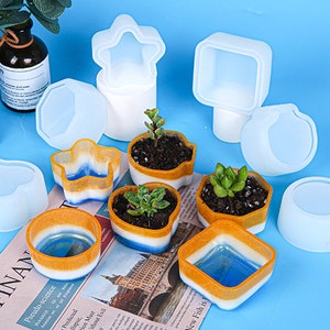 May include: Assortment of 10 white silicone molds for making various shapes of planters. The molds are in different shapes, including a star, a square, a heart, a circle, and a flower pot. The molds are on a blue surface with small plants in some of the molds.