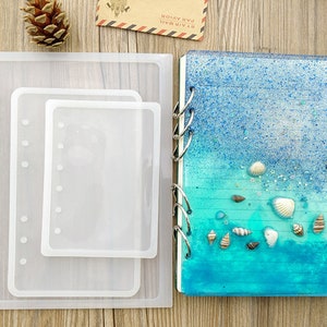 May include: A clear silicone mould for making resin crafts. The mould is shaped like a notebook with a spine and holes for binding. The mould is shown with a finished resin notebook with a blue and green ocean-themed design.
