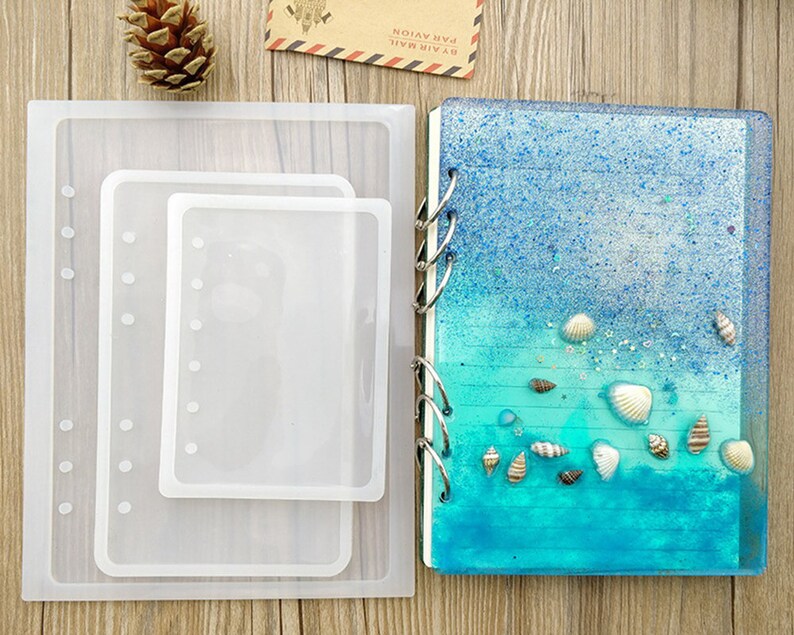 May include: A clear silicone mould for making resin crafts. The mould is shaped like a notebook with a spine and holes for binding. The mould is shown with a finished resin notebook with a blue and green ocean-themed design.