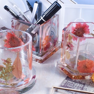 May include: Three clear resin pencil holders with dried flowers and leaves embedded inside. The holders are in different shapes, including a square, a cylinder, and a hexagon. The holders are sitting on a table with a piece of paper with music notes printed on it.