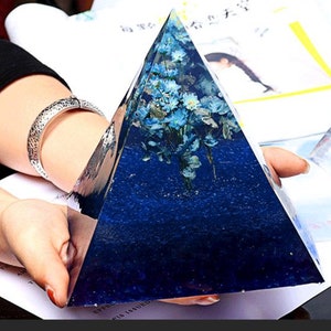 Peut inclure: Une pyramide en verre bleu avec des fleurs séchées à l'intérieur. La pyramide est transparente et a une surface texturée. Les fleurs sont bleues et blanches.