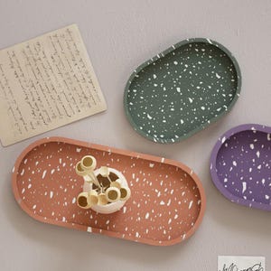 May include: Four oval-shaped decorative trays in shades of terracotta, green, and purple, speckled with white. One tray holds a small white vase with decorative elements. A handwritten note is in the background.