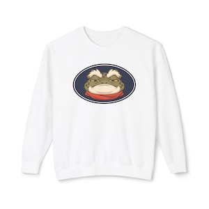 May include: A white long-sleeve sweatshirt featuring a centered graphic of a grumpy-looking toad with white eyebrows and a red scarf, enclosed within a dark blue oval frame. The toad has a textured green skin and yellow eyes.
