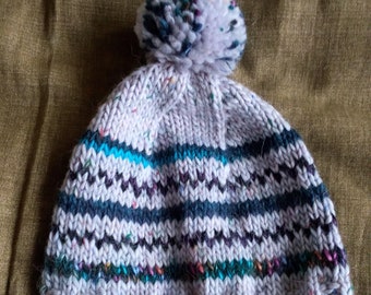 Puede incluir: Un gorro de punto con un pompón en la parte superior. El gorro está rayado en tonos de morado, azul y blanco. El gorro está hecho con un hilo jaspeado.