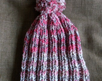 Puede incluir: Un gorro de punto con un pompón en la parte superior. El gorro está hecho con un hilo multicolor en tonos rosa, blanco y marrón.
