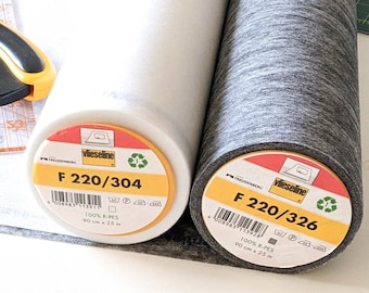 Peut inclure: Deux rouleaux d'entoilage Vlieseline, un blanc étiqueté F 220/304 et un gris anthracite étiqueté F 220/326, mesurant chacun 90 cm sur 25 m. Un cutter rotatif jaune et noir repose sur une règle de patchwork en plastique transparent à lignes orange.