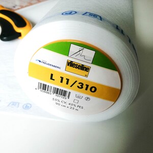 May include: A white roll of vlieseline interfacing with a yellow and green label. The label reads "vlieseline", "FREUDENBERG", "L 11/310", "4 008983 101123", "60P", "55% CV, 45% PES", and "90 cm x 25 m".