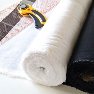 May include: A close-up of sewing equipment. Two fabric rolls, one white and one black, are next to a clear ruler and a yellow and black rotary cutter. The fabric is tightly rolled, and the cutter is open.