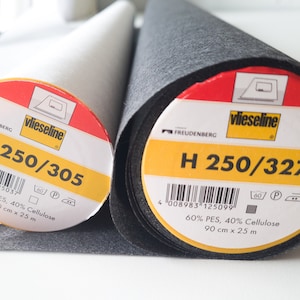 Könnte beinhalten: Zwei Rollen Vlieseline-Vlieseline, eine weiße und eine schwarze. Die Etiketten sind gelb mit schwarzem Text und zeigen den Produktcode H 250/305 und H 250/327. Die Etiketten geben auch die Materialzusammensetzung an: 60 % PES, 40 % Cellulose. Jede Rolle ist 90 cm x 25 m groß.