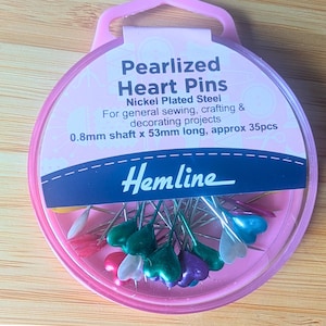 Könnte beinhalten: Eine rosa Dose mit perlmuttartigen Herzstiften der Marke Hemline. Die Packung enthält vernickelte Stahlstifte, 0,8 mm Schaft x 53 mm lang, zum Nähen, Basteln und Dekorieren. Die herzförmigen Stiftköpfe sind in verschiedenen Farben gehalten.