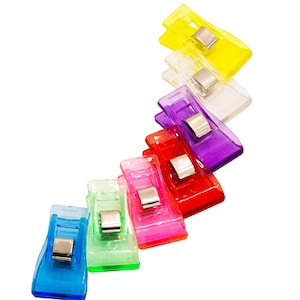 May include: A collection of colorful sewing clips in various shades, including blue, green, pink, red, purple, yellow, and clear. Each clip has a silver metal clasp. These plastic clips are designed for fabric and craft projects.