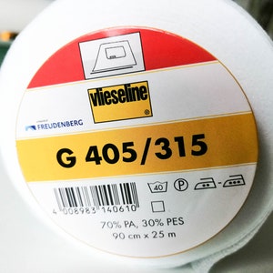 Peut inclure: Un rouleau de tissu blanc avec une étiquette portant l'inscription "Vlieseline G 405/315". L'étiquette comprend également le logo Freudenberg, des symboles d'entretien et les dimensions de 90 cm x 25 m.