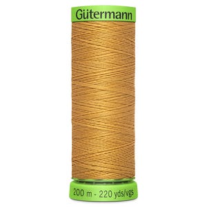 Könnte beinhalten: Eine Spule Gütermann-Garn in goldgelber Farbe. Die Spule hat grüne Enden mit dem Markennamen und den Garnmaßen aufgedruckt. Der Faden ist sauber um die Spule gewickelt.