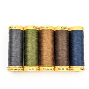 May include: Five spools of Gutermann thread in various shades of gray, green, brown, and blue. Each spool has a yellow label with black text. The thread is tightly wound, and the spools are arranged in a row.