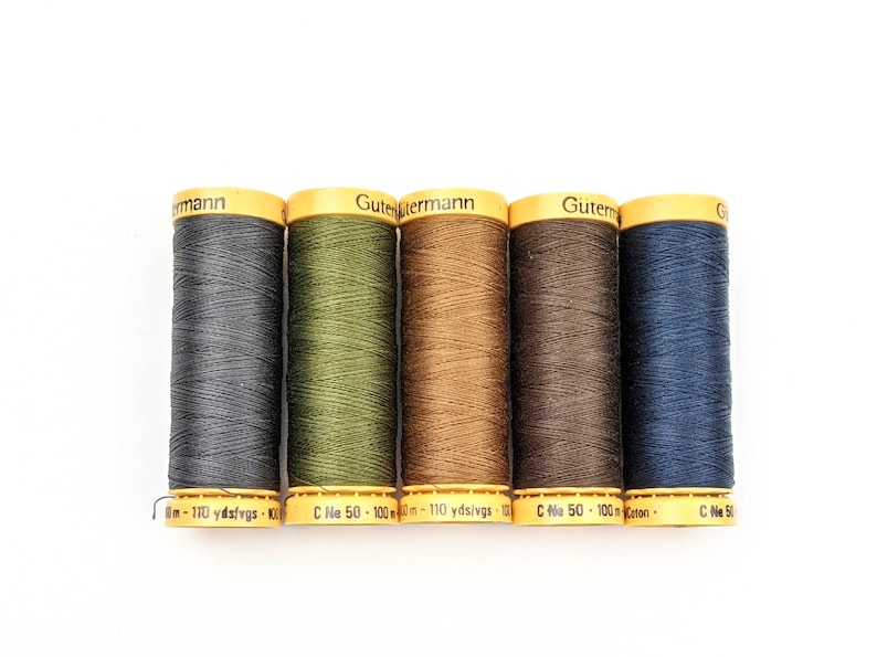 May include: Five spools of Gutermann thread in various shades of gray, green, brown, and blue. Each spool has a yellow label with black text. The thread is tightly wound, and the spools are arranged in a row.