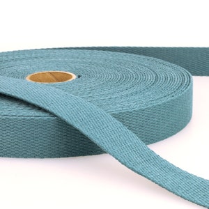May include: A roll of teal cotton webbing. The webbing is tightly woven and unwinds from a cardboard core. The colour is a muted, dusty teal, and the texture is slightly rough. The width of the webbing is approximately 2.5 cm.