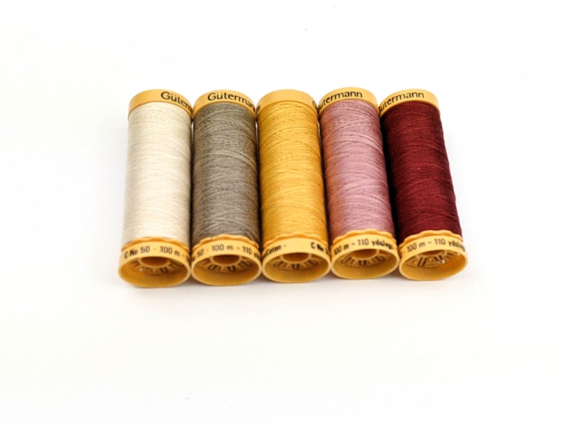 May include: Five spools of Gutermann thread in various colors: off-white, gray, mustard yellow, mauve, and burgundy. Each spool has a yellow base and a label with the brand name and thread length.