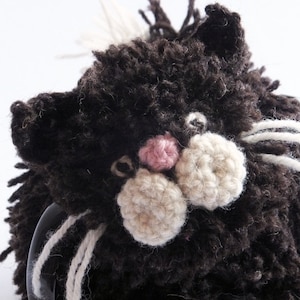 May include: A black and white crocheted cat mug cosy with a pink nose. The cosy has long white yarn whiskers.