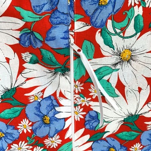 May include: A fabric textile featuring a vibrant red background with a dense floral pattern of large white daisies, blue flowers, and green leaves. A vertical white zip runs down the centre, equipped with a long white fabric pull tab.