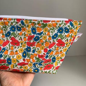 Può includere: Una pochette bianca con cerniera con una stampa floreale con papaveri rossi, margherite blu, campanelle gialle e foglie verdi.