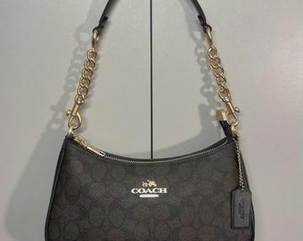 May include: A dark brown Coach shoulder bag featuring the signature monogram pattern, a gold-tone chain strap, a top zipper closure, and a matching leather hangtag. The front displays the gold Coach logo and horse and carriage emblem.