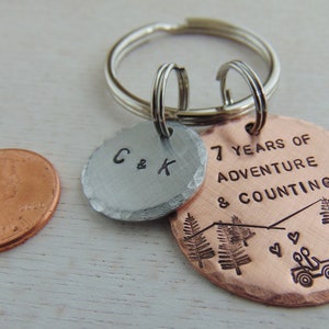 Puede incluir: Un llavero de cobre y plata con el texto "7 Years of Adventure & Counting" y dos &aacute;rboles en el dije de cobre. El dije de plata tiene las iniciales "C & K".