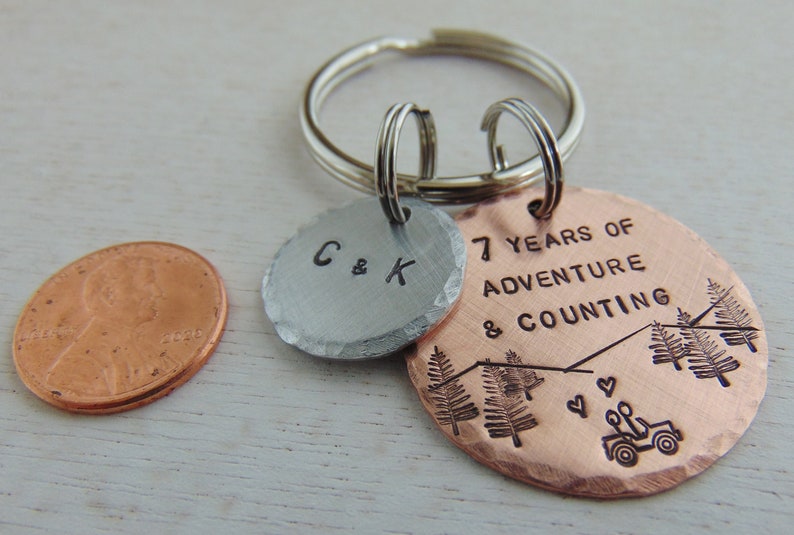 Puede incluir: Un llavero de cobre y plata con el texto "7 Years of Adventure & Counting" y dos &aacute;rboles en el dije de cobre. El dije de plata tiene las iniciales "C & K".