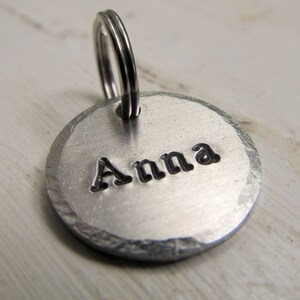 K&ouml;nnte beinhalten: Silberner Hundeanh&auml;nger aus Metall mit abgerundeter Kante und einem geteilten Ring. Der Anh&auml;nger ist mit dem Namen "Anna" graviert.