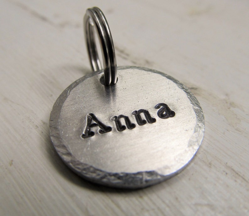 K&ouml;nnte beinhalten: Silberner Hundeanh&auml;nger aus Metall mit abgerundeter Kante und einem geteilten Ring. Der Anh&auml;nger ist mit dem Namen "Anna" graviert.