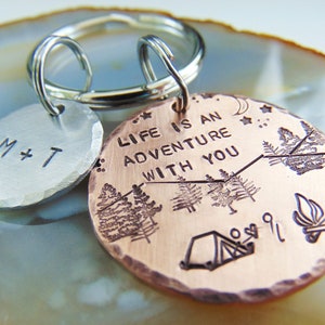 May include: A copper keychain with the text "Life is an adventure with you" engraved on it. The keychain also features a small silver tag with the initials "M+T" engraved on it. The copper tag has a design of a tent, a campfire, and trees.