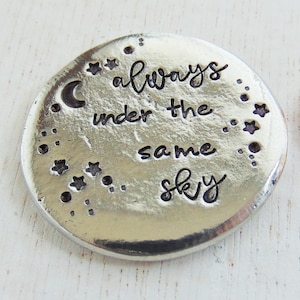 May include: A silver round charm with the text "always under the same sky" and a crescent moon and stars around the text.