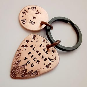 May include: A copper-colored keychain with a guitar pick-shaped charm that reads "I'll Always Pick You." The keychain also includes a round charm with initials and a date. The charms are connected to a black key ring.