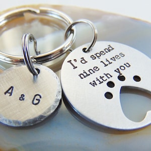 May include: A silver keychain with two round pendants. One pendant has the initials "A & G" stamped on it. The other pendant has the phrase "I'd spend nine lives with you" stamped on it, with a heart-shaped cutout.