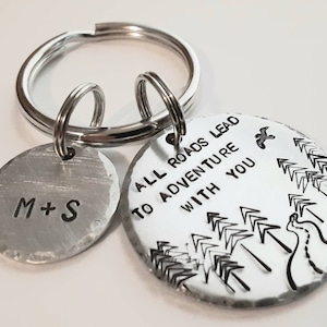 May include: Silver keychain with two round tags. One tag has the text "M+S" and the other tag has the text "All roads lead to adventure with you" and a design of trees and a winding path.
