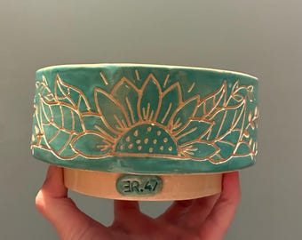 Op de afbeelding: Een groenblauwe keramieken kom met een uitgesneden zonnebloemmotief met goudkleurige omlijningen. De basis is van ongeglazuurd beige keramiek en bevat een klein ovaal label met de tekst ER.47 in reliëf in groenblauw op de voorzijde.