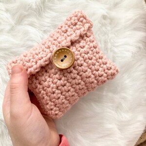 Peut inclure: Une pochette en crochet rose avec une fermeture bouton en bois. La pochette est tenue dans une main.