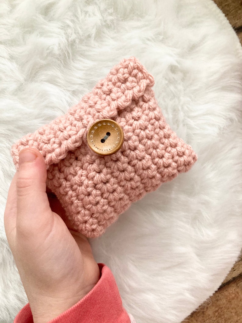 Peut inclure: Une pochette en crochet rose avec une fermeture bouton en bois. La pochette est tenue dans une main.