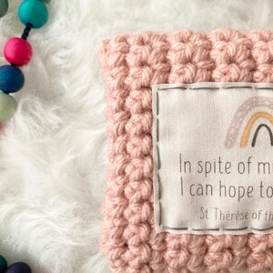 Peut inclure: Un coussin en crochet rose avec une &eacute;tiquette en tissu blanc qui dit "Malgr&eacute; ma faiblesse, j'esp&egrave;re faire de grandes choses. Sainte Th&eacute;r&egrave;se de l'Enfant J&eacute;sus".