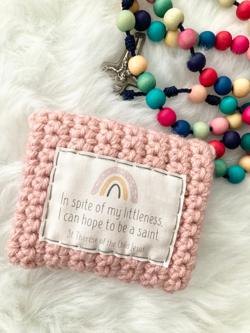 Peut inclure: Une pochette en crochet rose avec un patch en tissu blanc repr&eacute;sentant un arc-en-ciel et la citation "Malgr&eacute; ma petitesse, j'esp&egrave;re &ecirc;tre sainte. Sainte Th&eacute;r&egrave;se de l'Enfant J&eacute;sus."