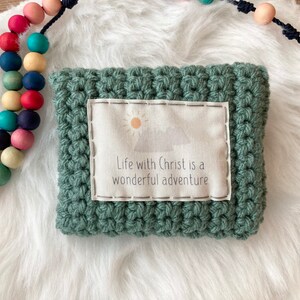 Peut inclure: Une pochette en crochet vert avec un insert en tissu blanc qui dit "Life with Christ is a wonderful adventure". La pochette est pos&eacute;e sur une surface blanche et moelleuse &agrave; c&ocirc;t&eacute; d'un chapelet en bois color&eacute;.