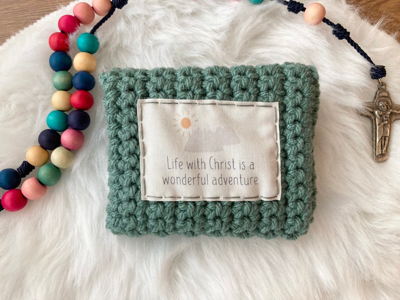 Peut inclure: Une pochette en crochet vert avec un insert en tissu blanc qui dit "Life with Christ is a wonderful adventure". La pochette est pos&eacute;e sur une surface blanche et moelleuse &agrave; c&ocirc;t&eacute; d'un chapelet en bois color&eacute;.