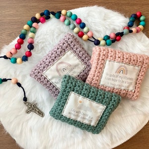 Peut inclure: Trois pochettes en crochet avec des citations inspirantes et des motifs arc-en-ciel. Les pochettes sont rose, violette et verte. Un chapelet en bois color&eacute; avec une croix en argent est sur une surface blanche et moelleuse.